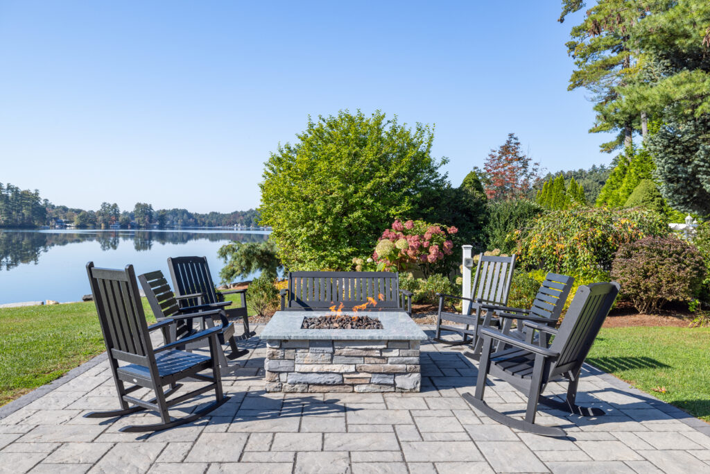 A set of patio chairs around a fireplace next to a lake and a garden