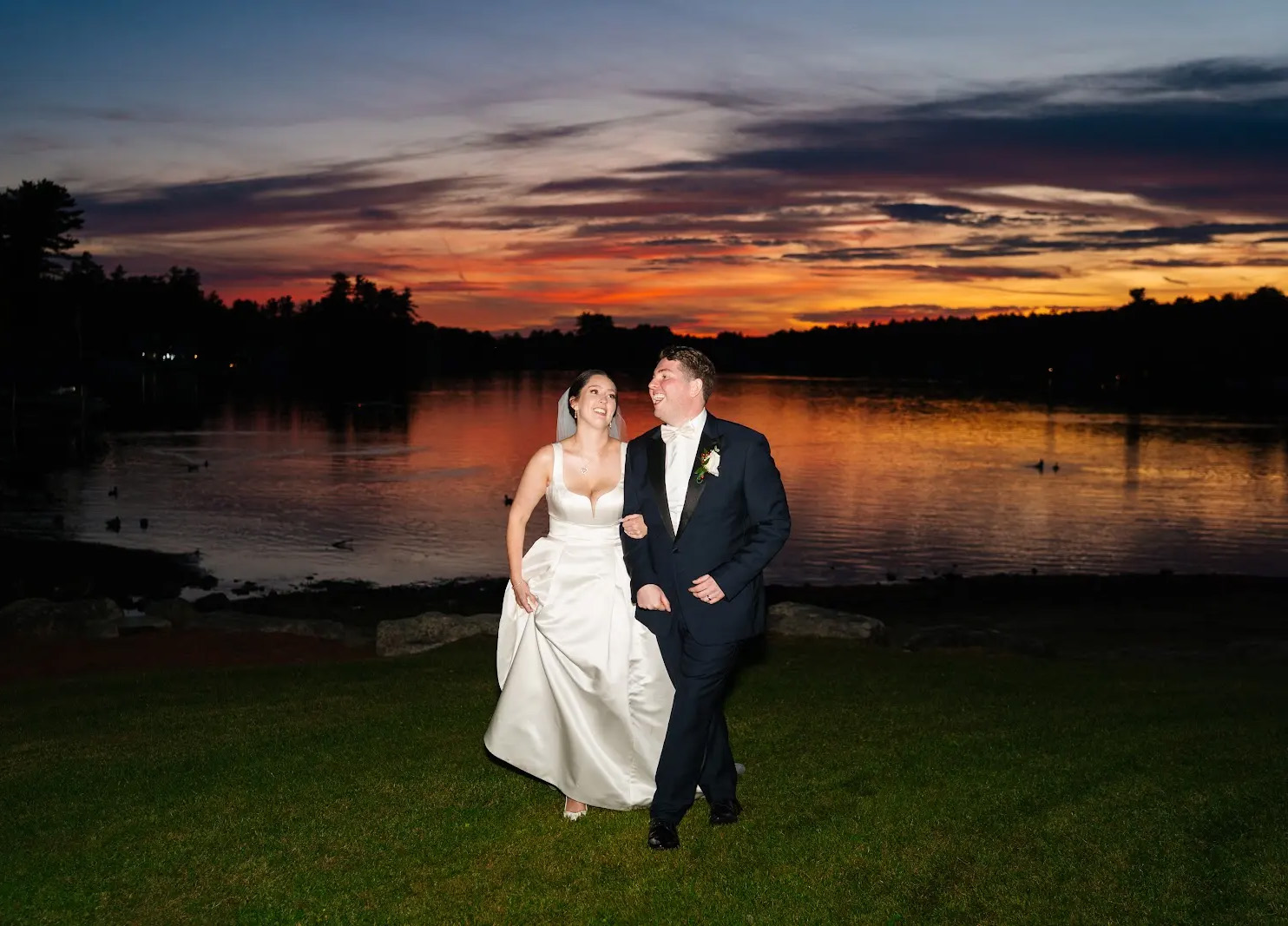 a couple stands in front of the lake at castleton in their wedding attire with a sunset in the background