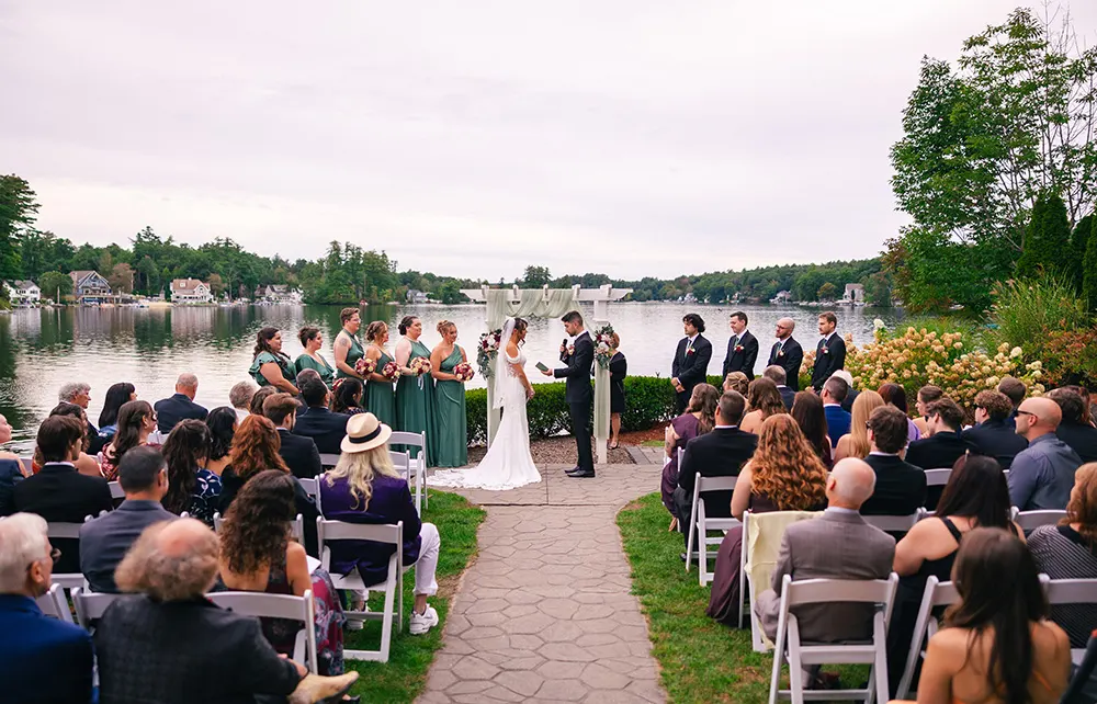 couple stands in front of the alter during an outdoor wedding in front of a lake with a crowd of people watching