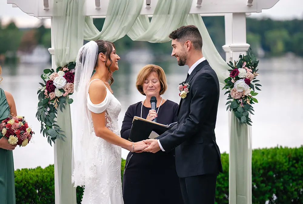 a young couple says their vows in front of landscaped shrubs with the lake in the background
